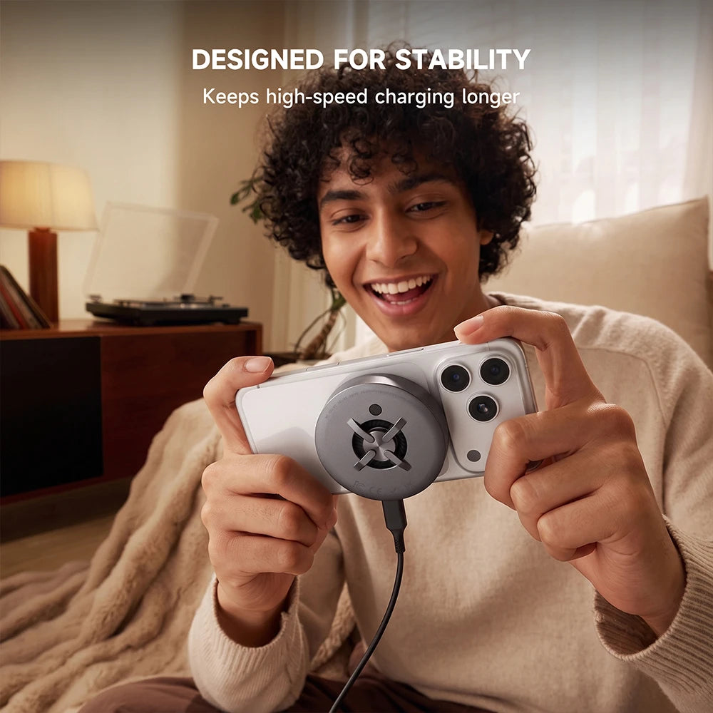 A smiling man in a beige sweater holds a white smartphone with a CoreDock Magnetic Wireless Charger attached to the back, seated in a warmly lit living room. Text at the top reads, "DESIGNED FOR STABILITY" and "Keeps high-speed charging longer." #color_gray