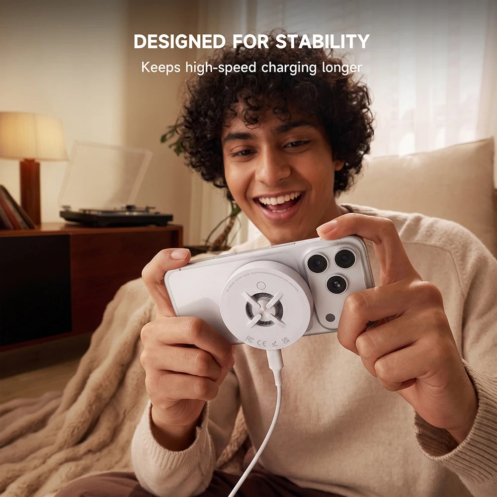 A smiling man in a cozy living room holding a white smartphone connected to a white CoreDock charging accessory, with top text reading “DESIGNED FOR STABILITY — Keeps high-speed charging longer.” #color_white