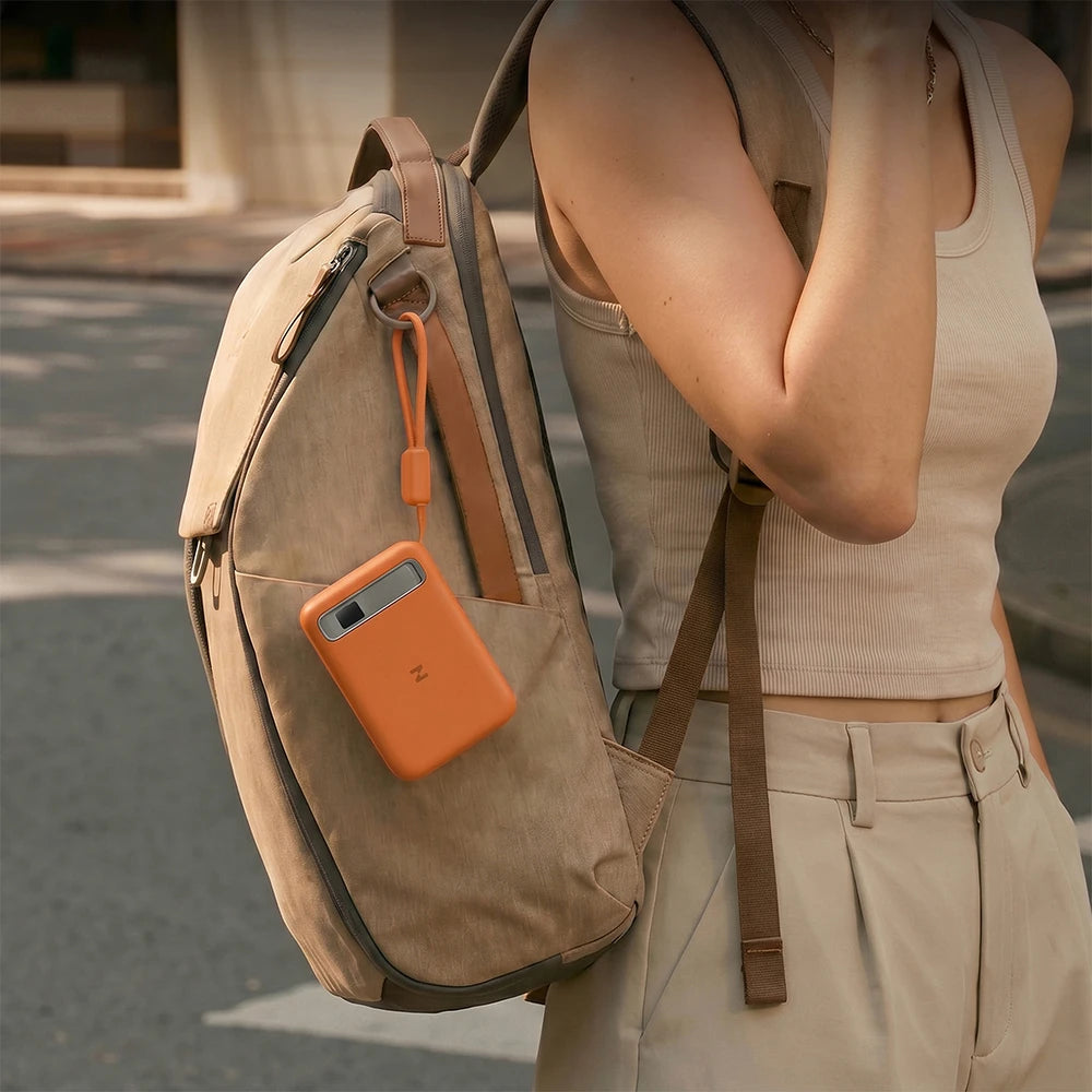 Person wearing a beige backpack with an orange MagAir magnetic wireless power bank attached, standing on a street.
