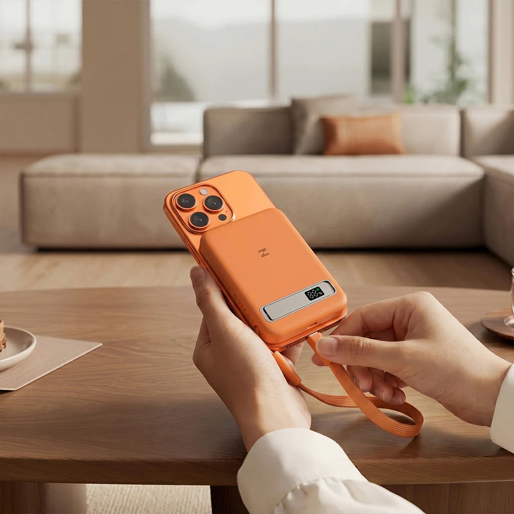 Person holding an orange smartphone with a matching orange MagAir magnetic wireless power bank in a living room setting.